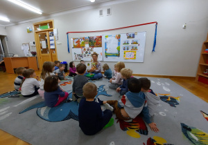 Dzieci siedzą na dywanie, a mama Tomka pokazuje im ilustrację do czytanego opowiadania.